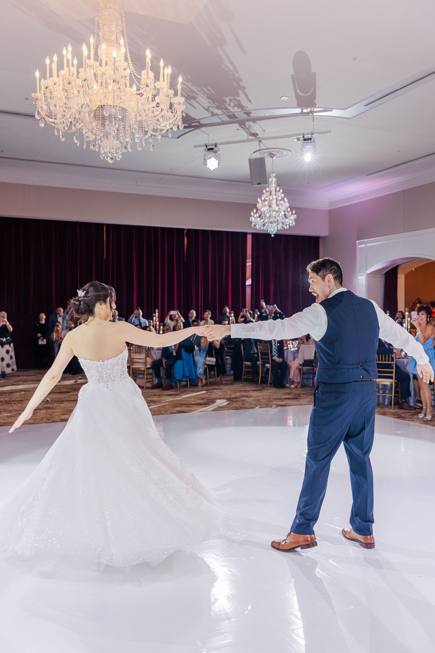 Bride and groom share their first dance under crystal chandeliers in an elegant ballroom with guests watching.