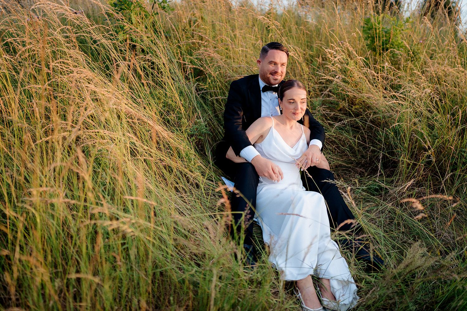 A couple sits on the ground together among tall wild grasses during a romantic golden hour photoshoot.
