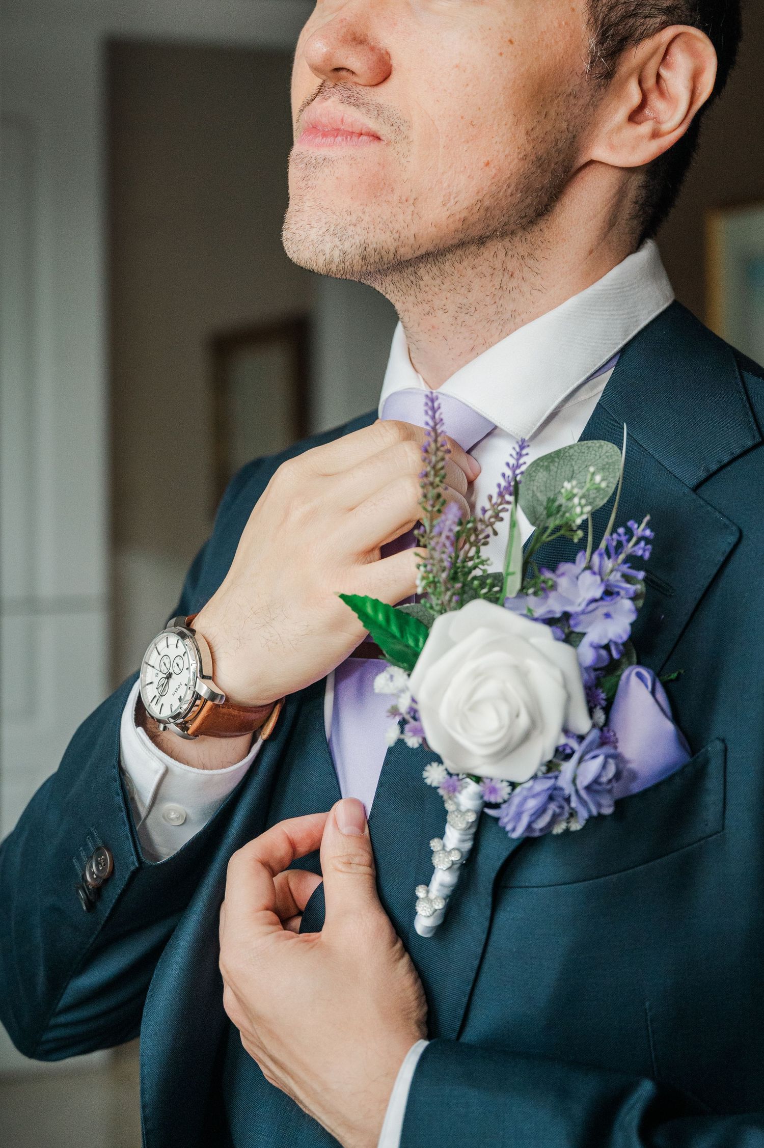 Groom in navy suit adjusting lavender tie with white rose and purple floral boutonniere, wearing a brown leather watch.