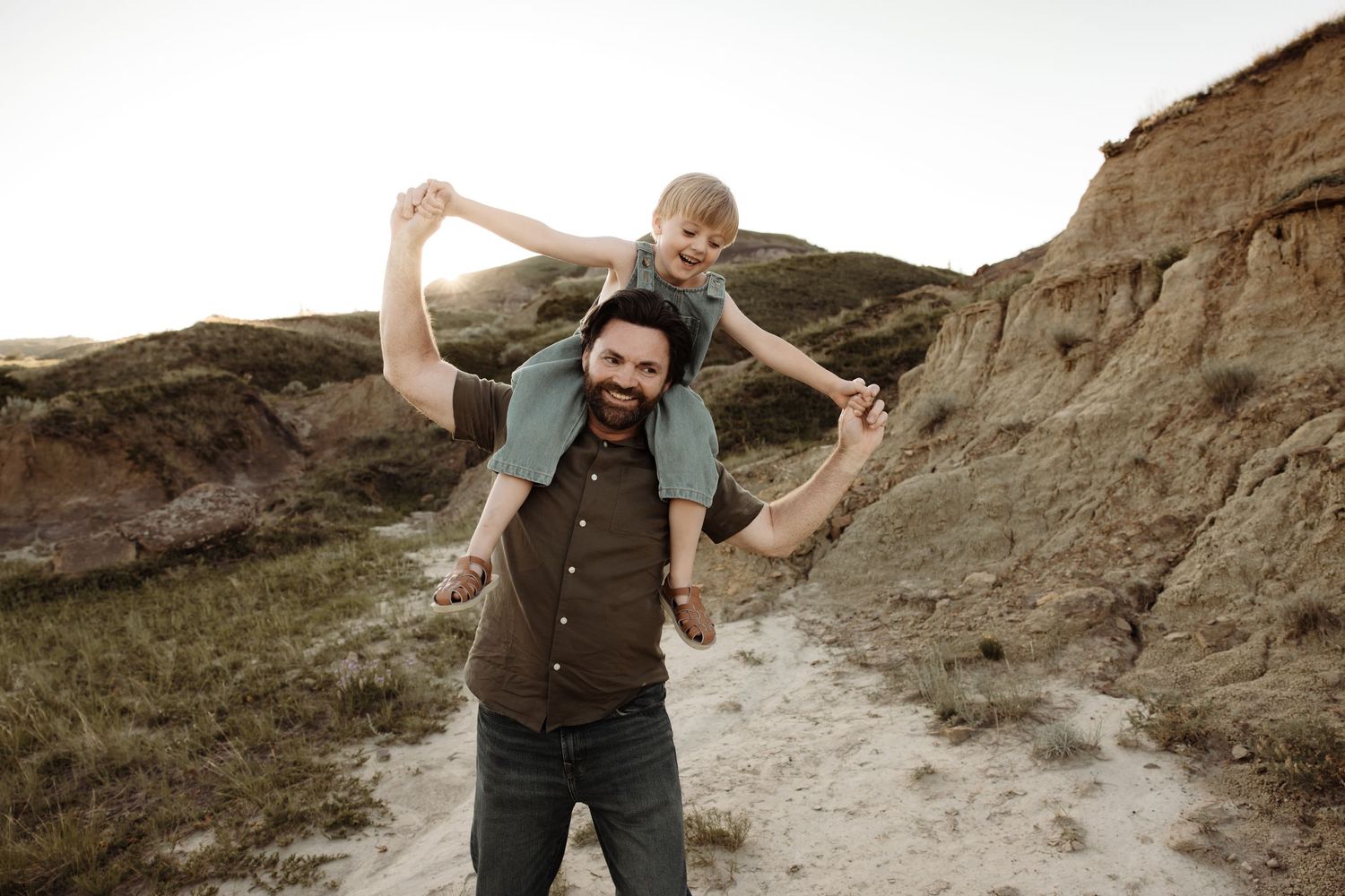 A bearded man carries a young child on his shoulders while hiking through a scenic sandy trail with rocky cliffs.