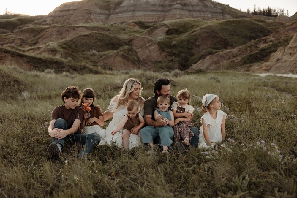 Family of seven sitting together in a grassy field with scenic badlands hills in the background as a photographer takes thier pictures