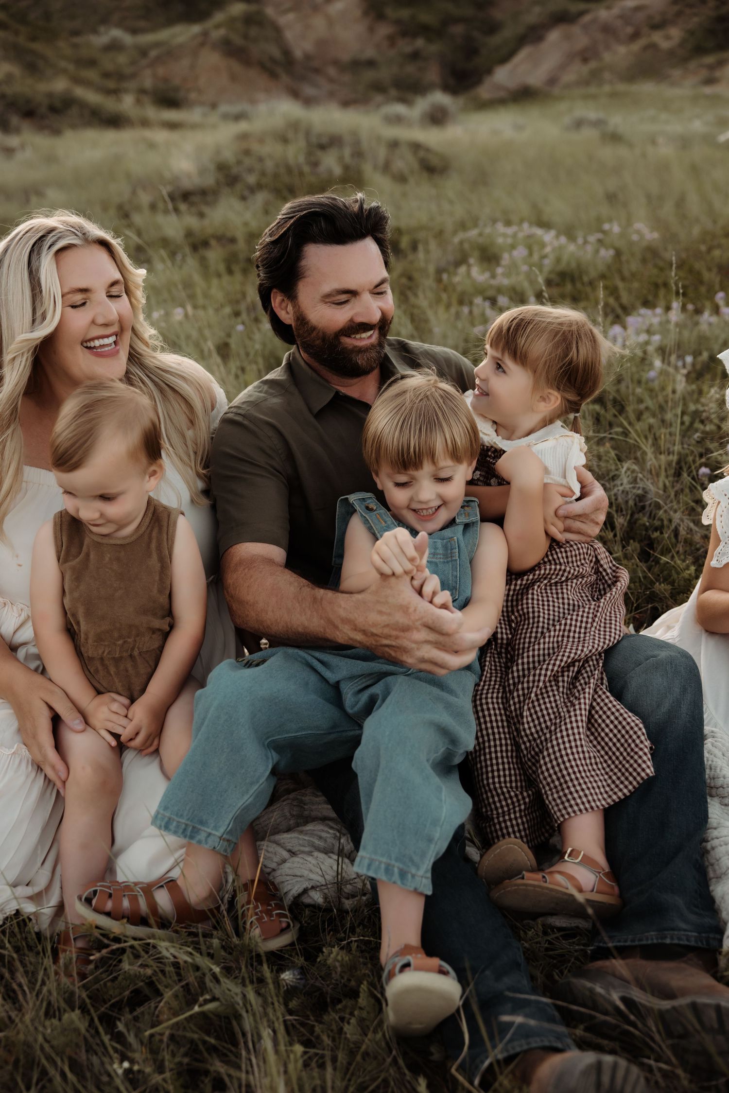 A joyful family of five sits together in a lush green meadow, parents laughing with three young children.
