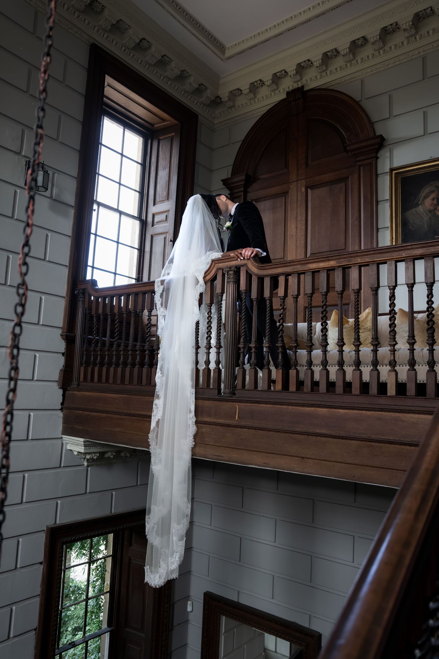 Bride and groom share a kiss on an elegant wooden balcony inside a historic stone building, bridal veil cascading down.