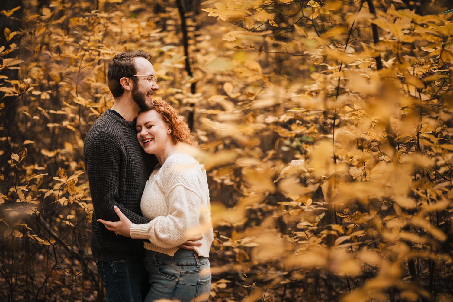 Engaged couple laughing softly, embraced closely in a dense autumn forest at Borderland State Park, framed by golden foliage.