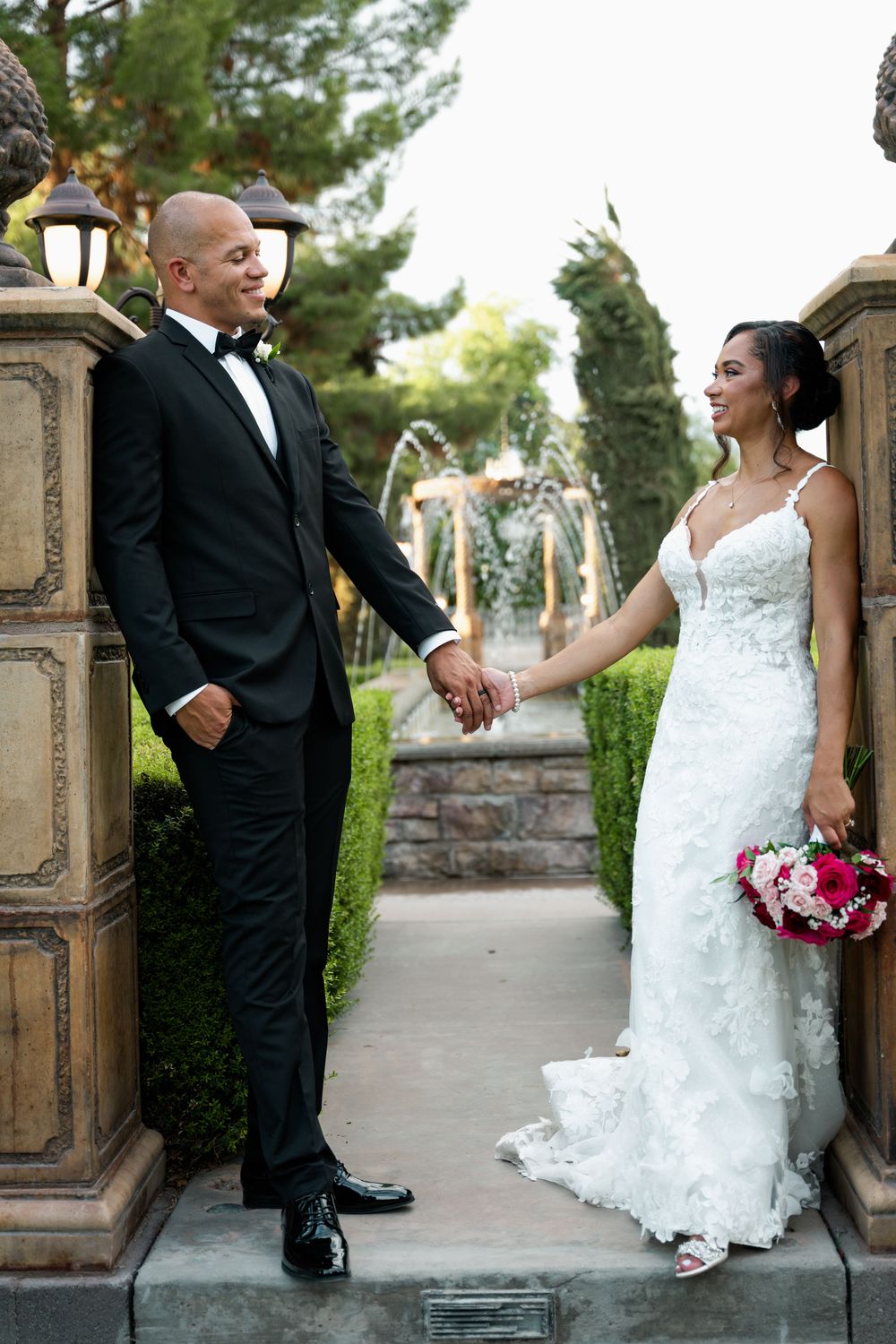 Bride and groom hold hands in elegant garden wedding venue with fountain backdrop, she in lace gown, he in black tuxedo.