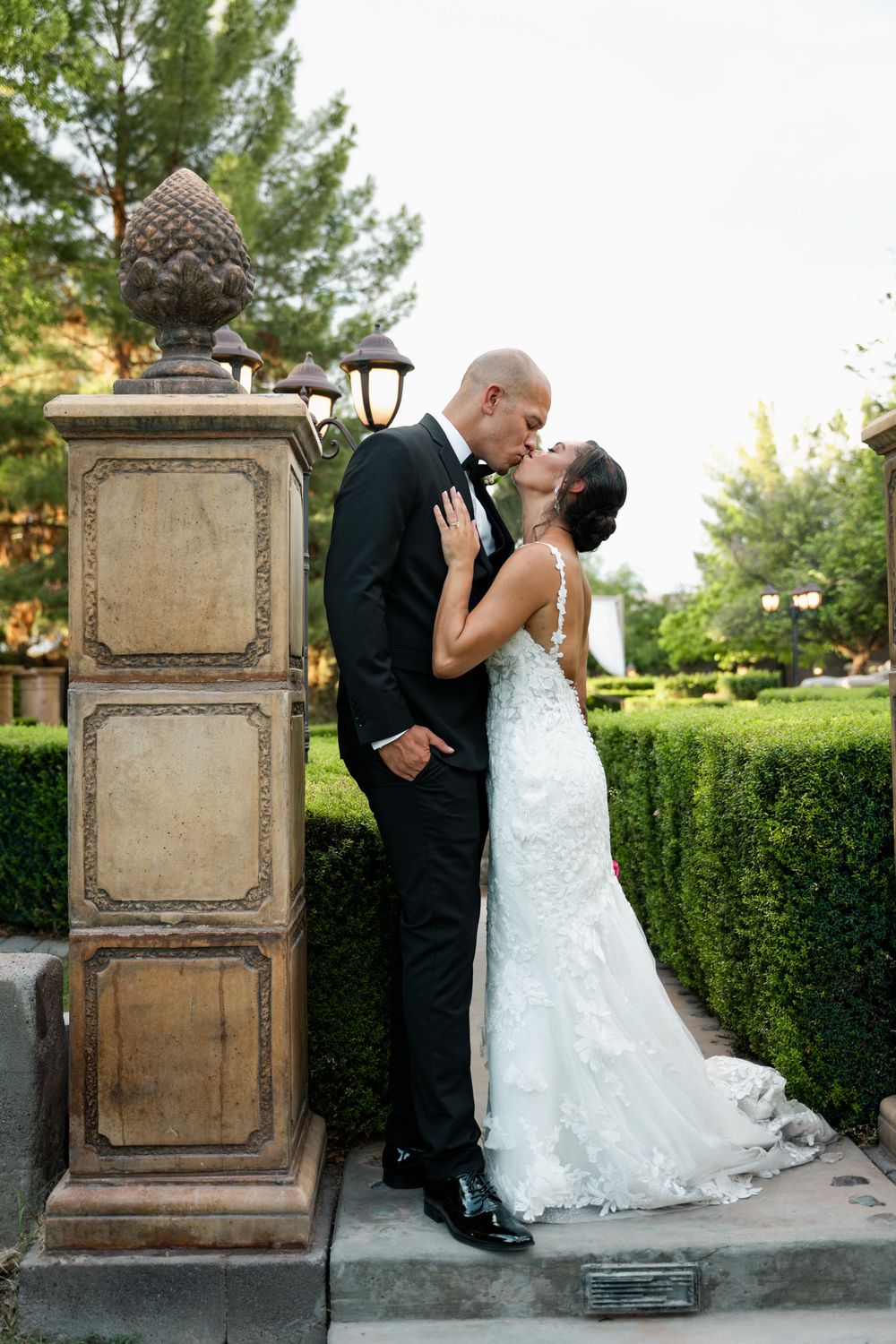 Bride and groom share a romantic kiss in a lush garden setting, with the bride in a lace gown and groom in a black tuxedo.