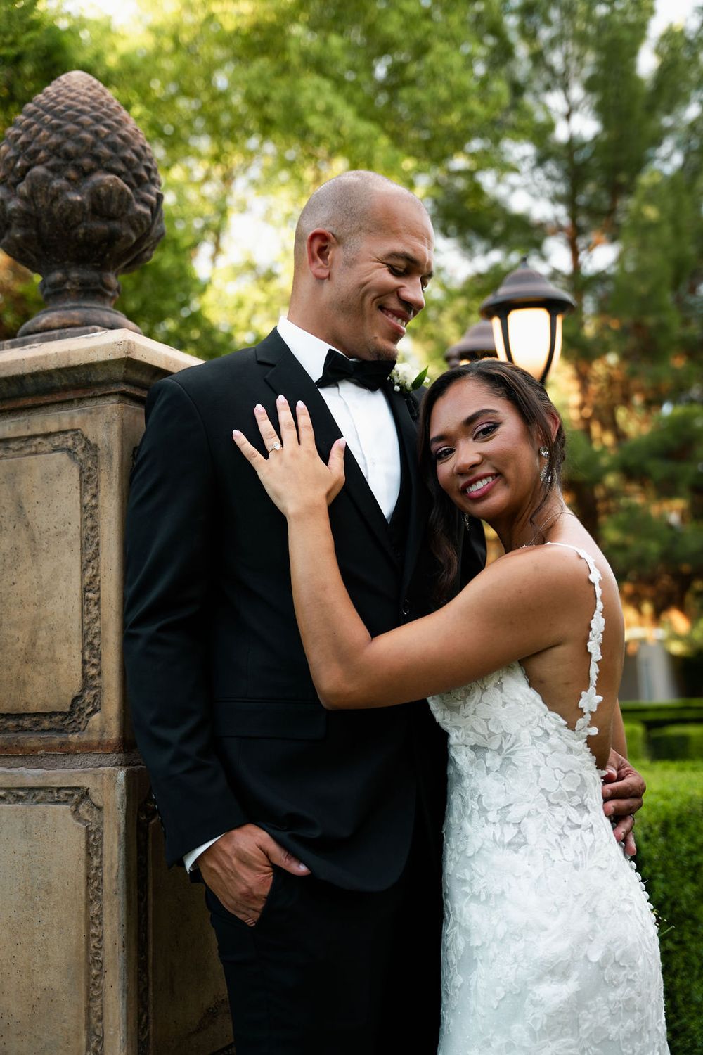 A smiling bride in a lace gown embraces her groom in a black tuxedo outdoors at a garden wedding venue.