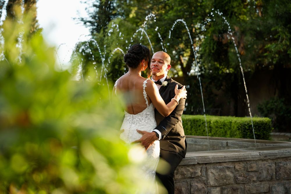 Bride and groom share romantic first dance near elegant fountain surrounded by lush greenery at outdoor wedding venue.