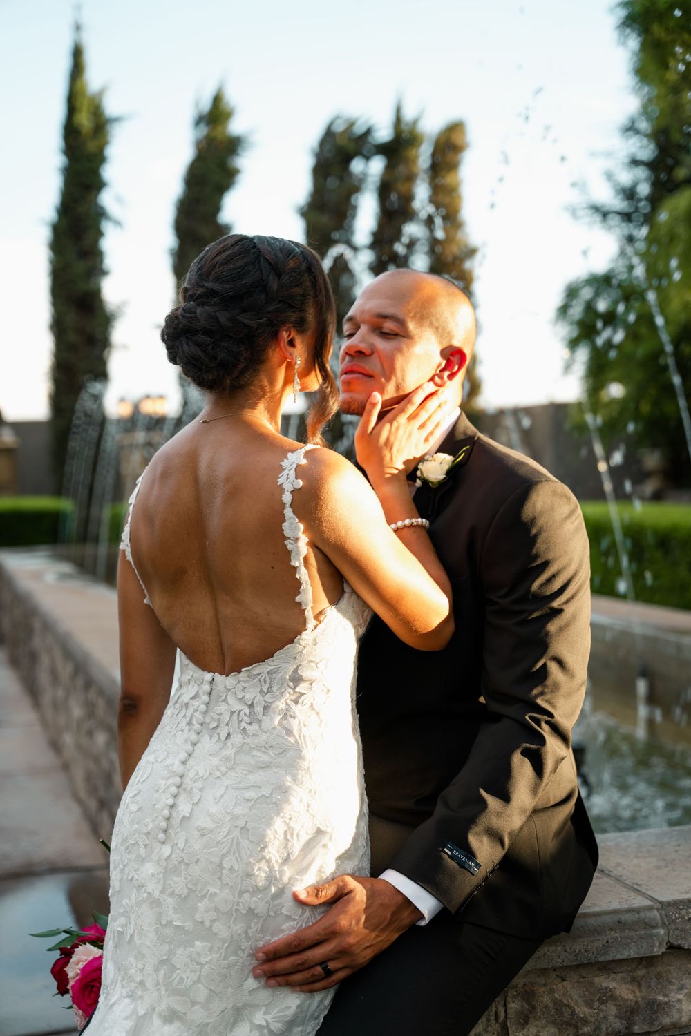 Bride in lace gown tenderly cups groom's face near fountain at outdoor wedding ceremony.
