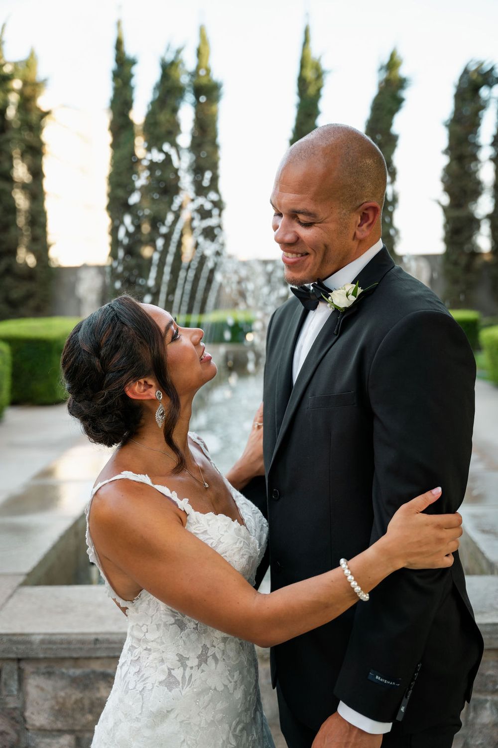 A bride and groom share a romantic embrace in front of a fountain, dressed in a white lace gown and black tuxedo.