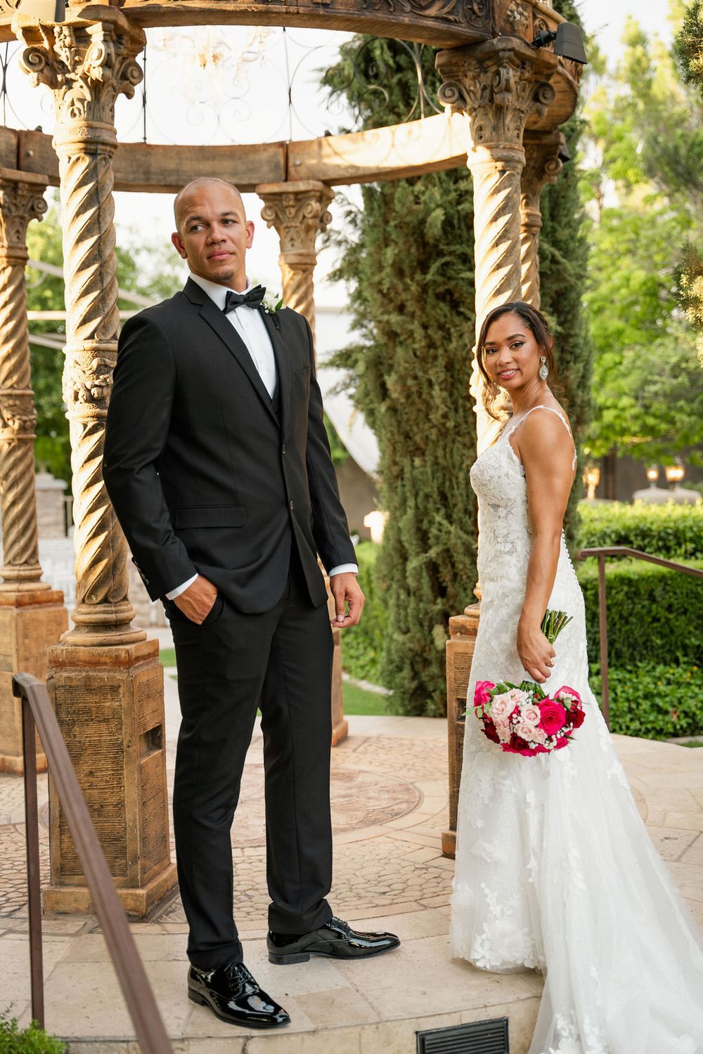Bride in lace gown holding pink bouquet stands with tuxedo-clad groom under ornate stone pergola outdoors.