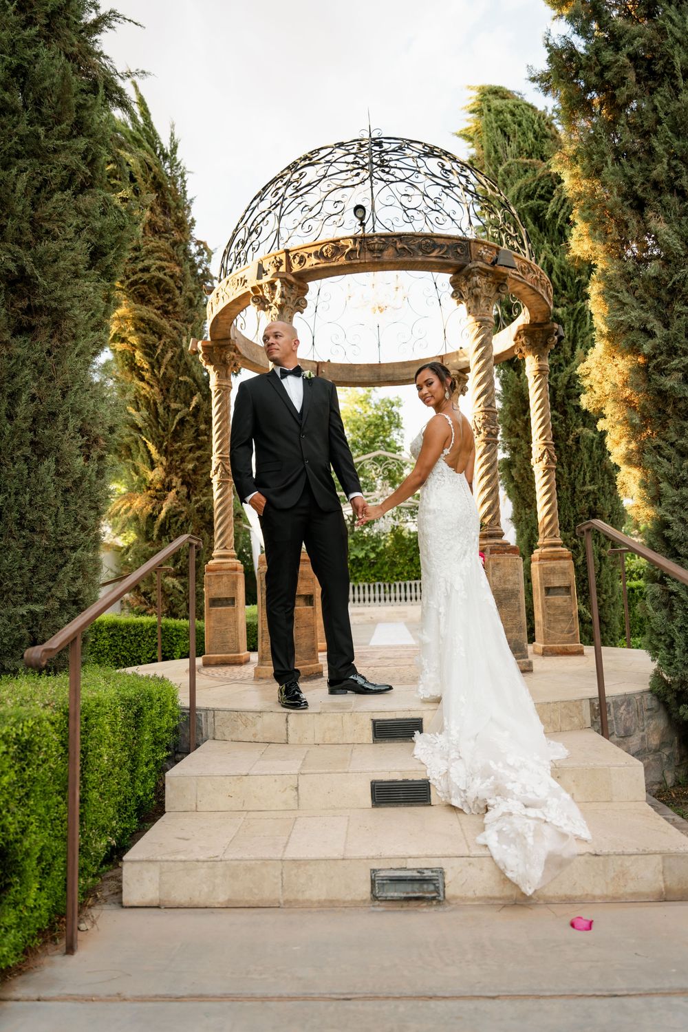 Bride and groom holding hands under ornate iron gazebo on stone steps surrounded by tall cypress trees.