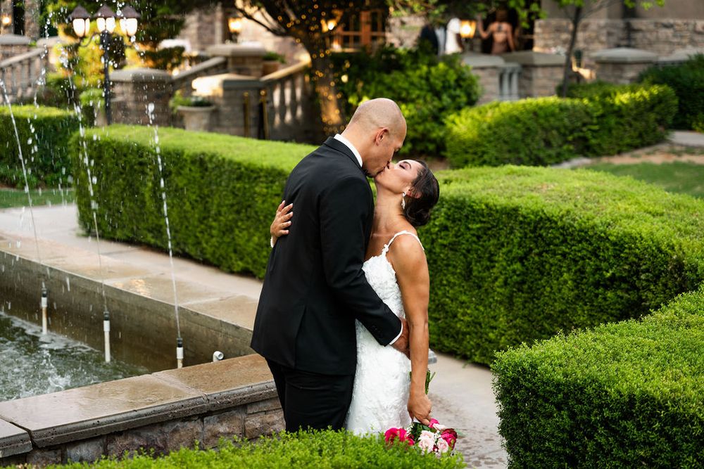 Bride and groom sharing a romantic kiss in a lush garden with fountains at their elegant outdoor wedding.