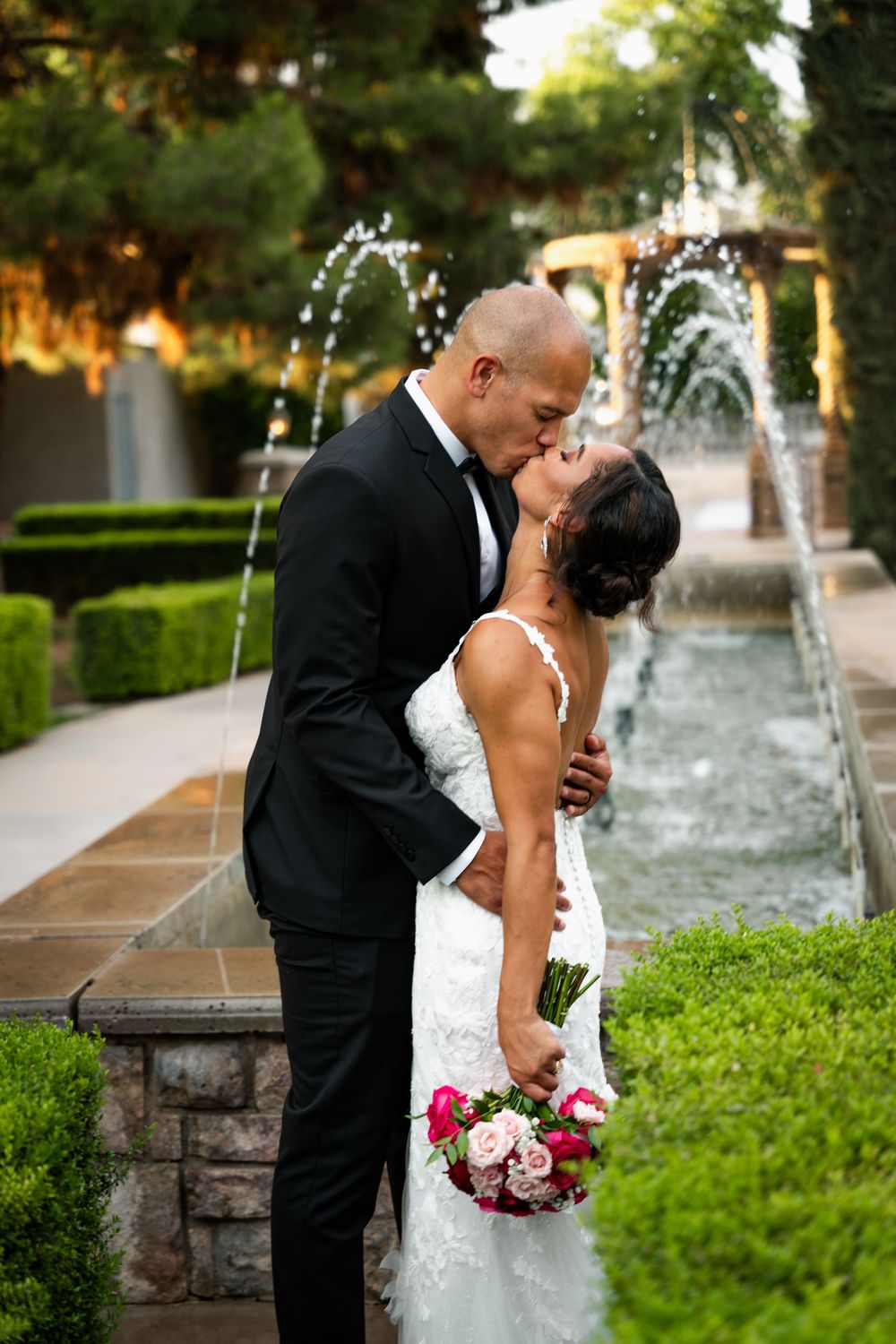 Bride and groom share a romantic kiss in front of a garden fountain, bride holding pink bouquet in white lace gown.