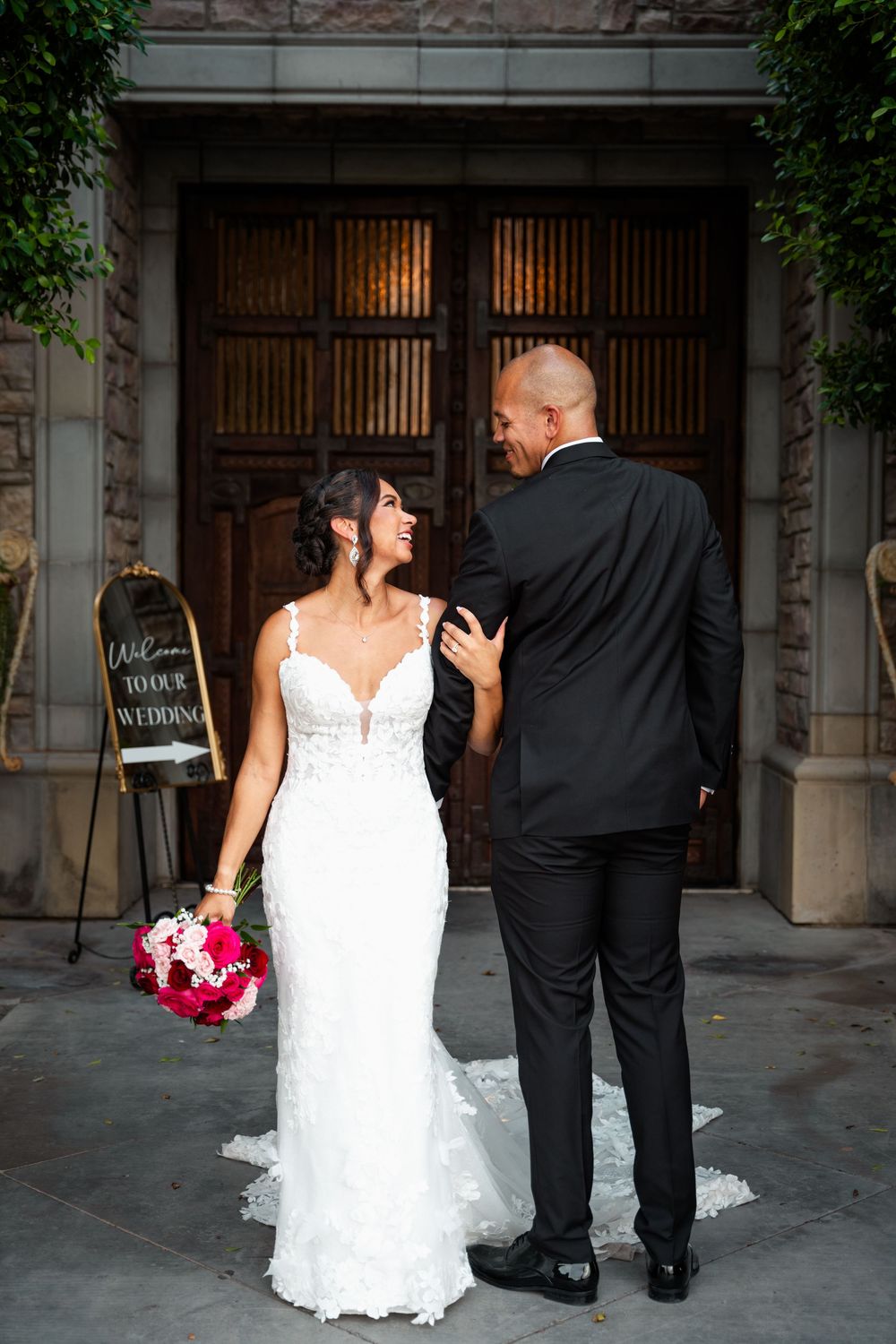 Bride in white lace gown holding pink bouquet smiles at groom in black suit before wooden doors with wedding sign.