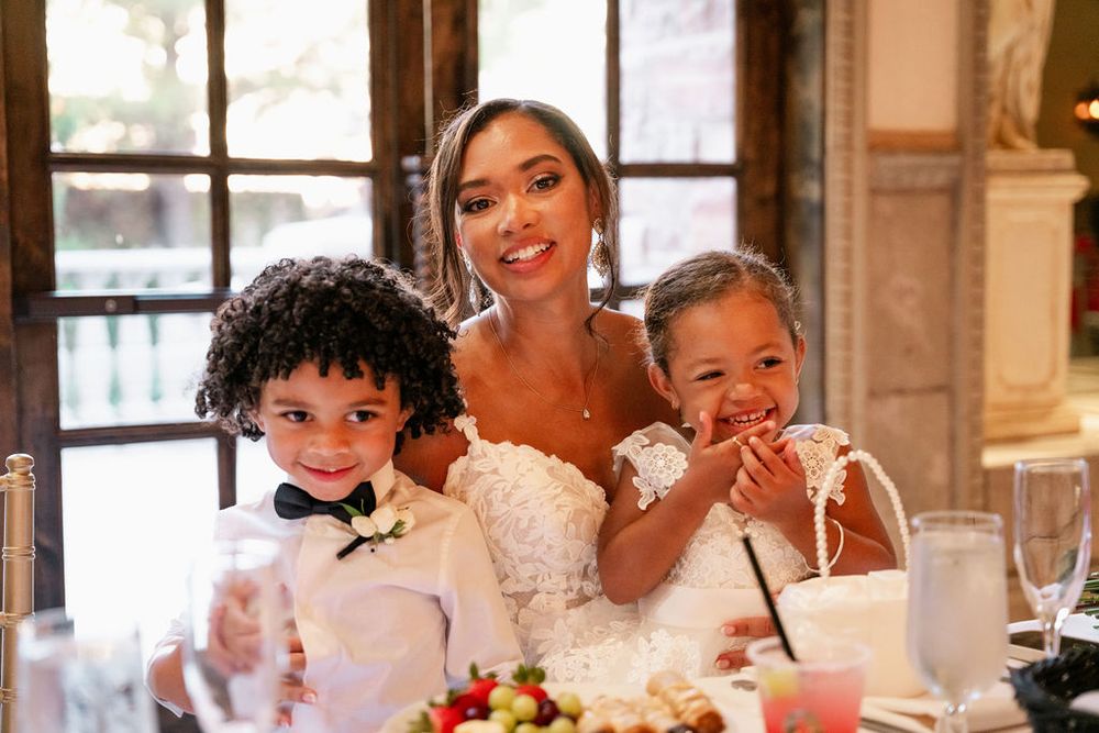 Bride in white lace gown smiling with two young children at a wedding reception table.