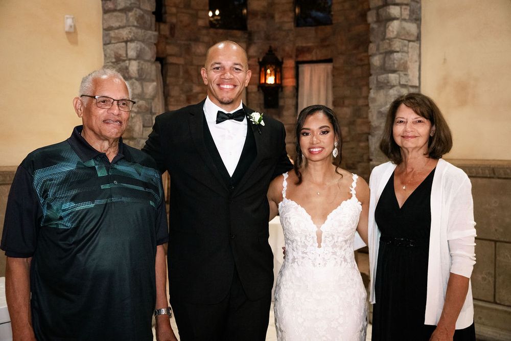 Wedding couple in formal attire poses with two family members inside stone venue.