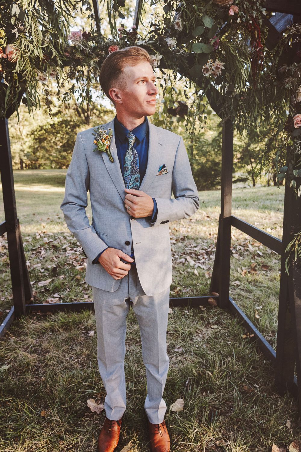 Groom in light gray suit with floral boutonniere and blue tie stands under decorated arch at outdoor wedding.