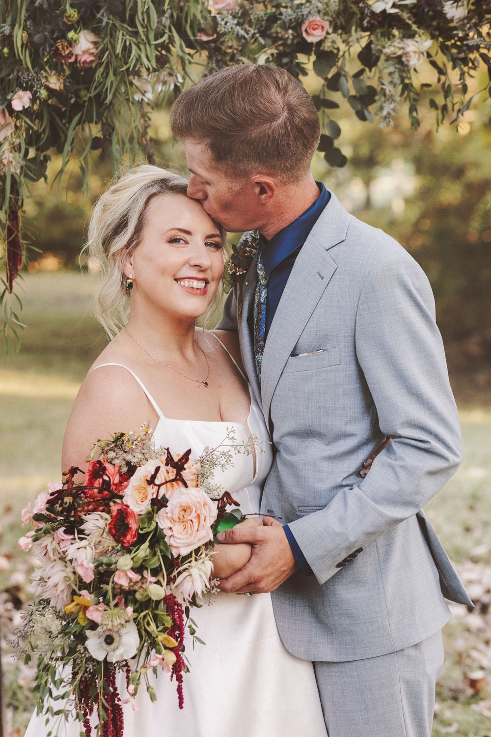 Bride smiling with colorful bouquet as groom kisses her cheek under floral arch at outdoor wedding.