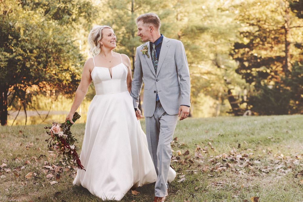 Bride and groom walking hand in hand outdoors in a sunlit garden, she in a white gown, he in a gray suit.