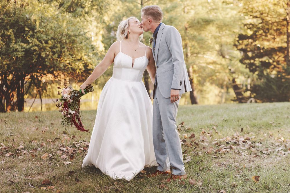 Bride and groom share a romantic kiss outdoors, surrounded by autumn foliage, bride holding a bouquet.