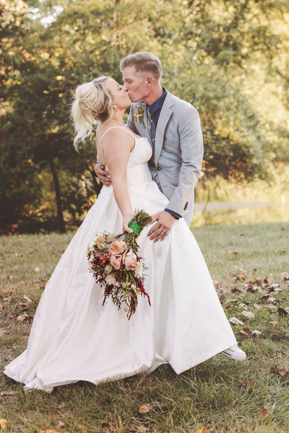 Bride and groom sharing a romantic kiss outdoors in autumn, bride holding a colorful bouquet in a white gown.