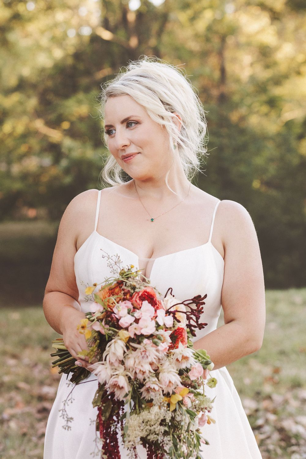 Blonde bride in white spaghetti strap gown holds cascading floral bouquet with pink and red blooms outdoors.