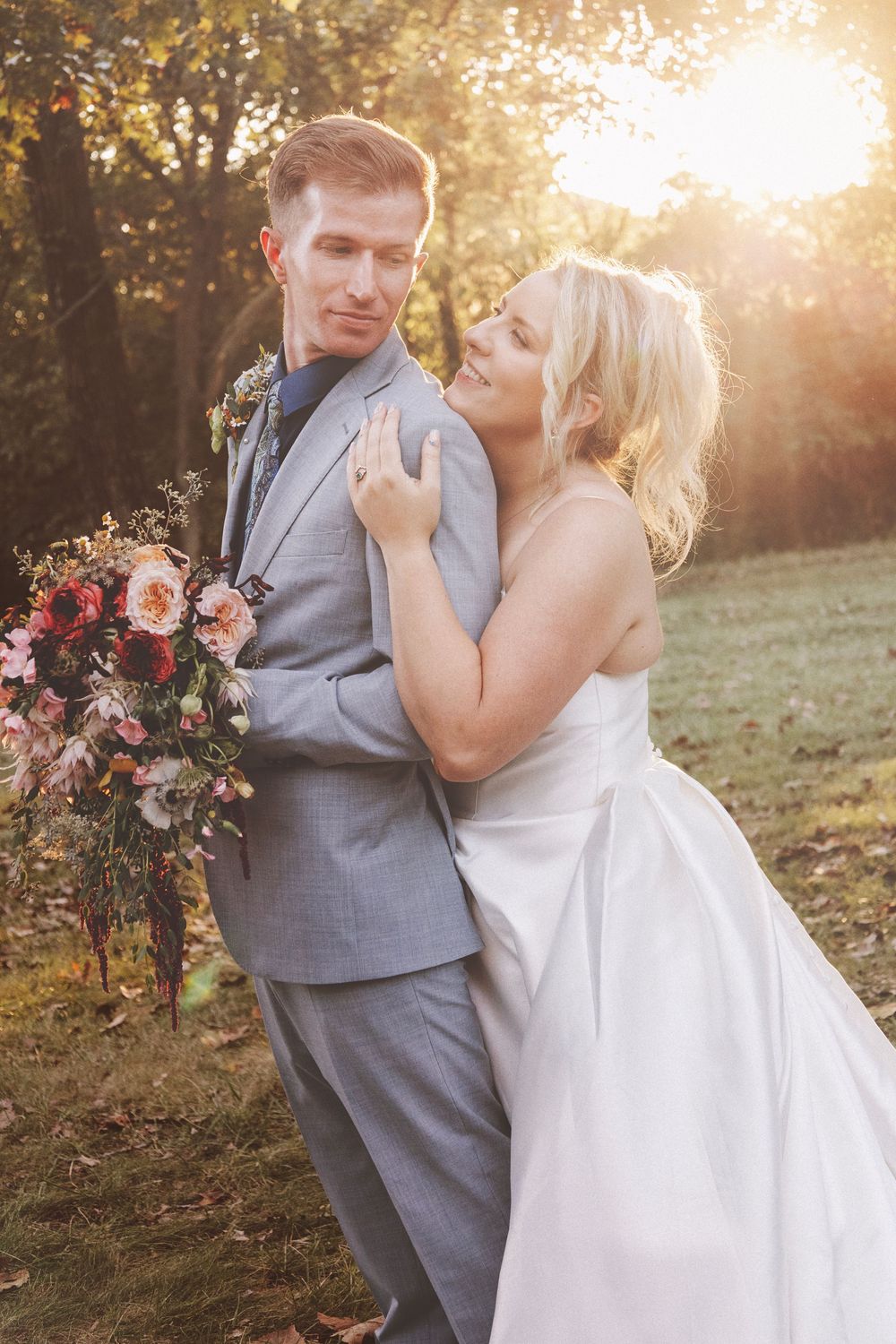 Bride in white gown embraces groom in gray suit outdoors, holding a lush floral bouquet in golden sunset light.