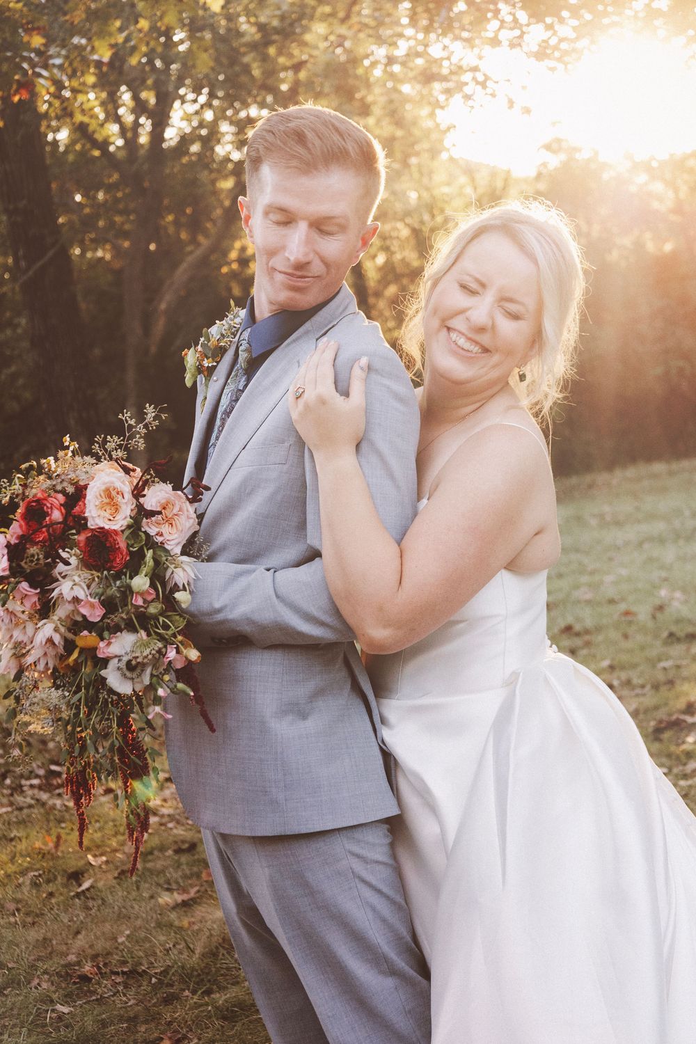 Bride and groom embrace outdoors at golden hour, she holds colorful bouquet in white gown, he wears gray suit.
