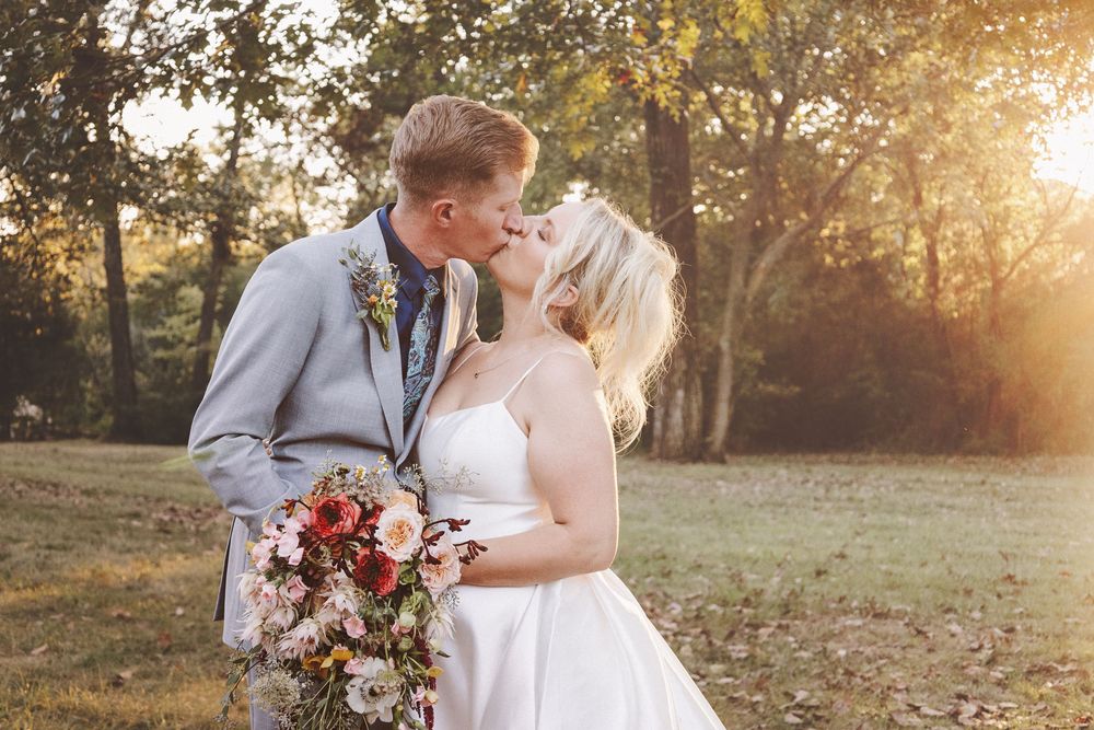 Bride and groom share a romantic kiss outdoors at golden hour, bride holding a colorful floral bouquet.