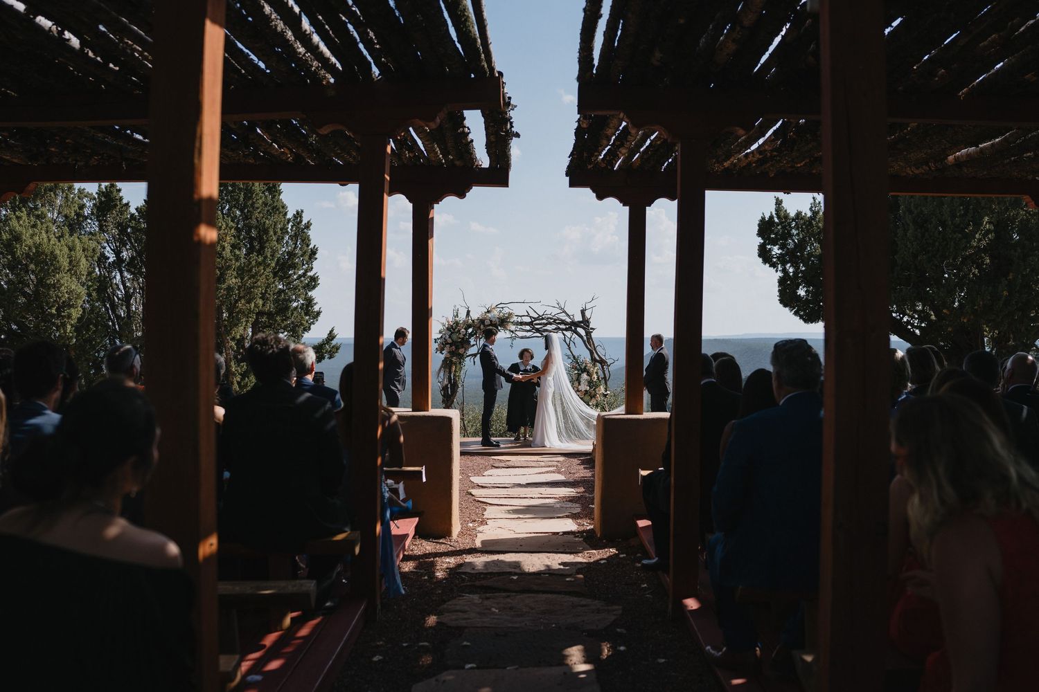 Outdoor wedding ceremony under rustic pergola with couple at floral arch, guests seated on both sides, mountain view backdrop.