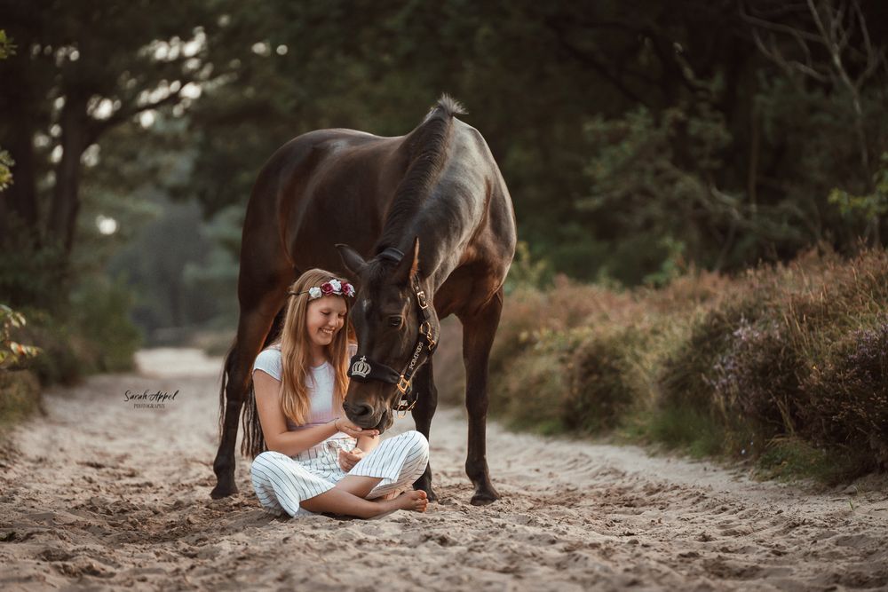 Girl with long hair and a flower crown sits on a sandy forest path as a dark bay horse lowers its head beside her.