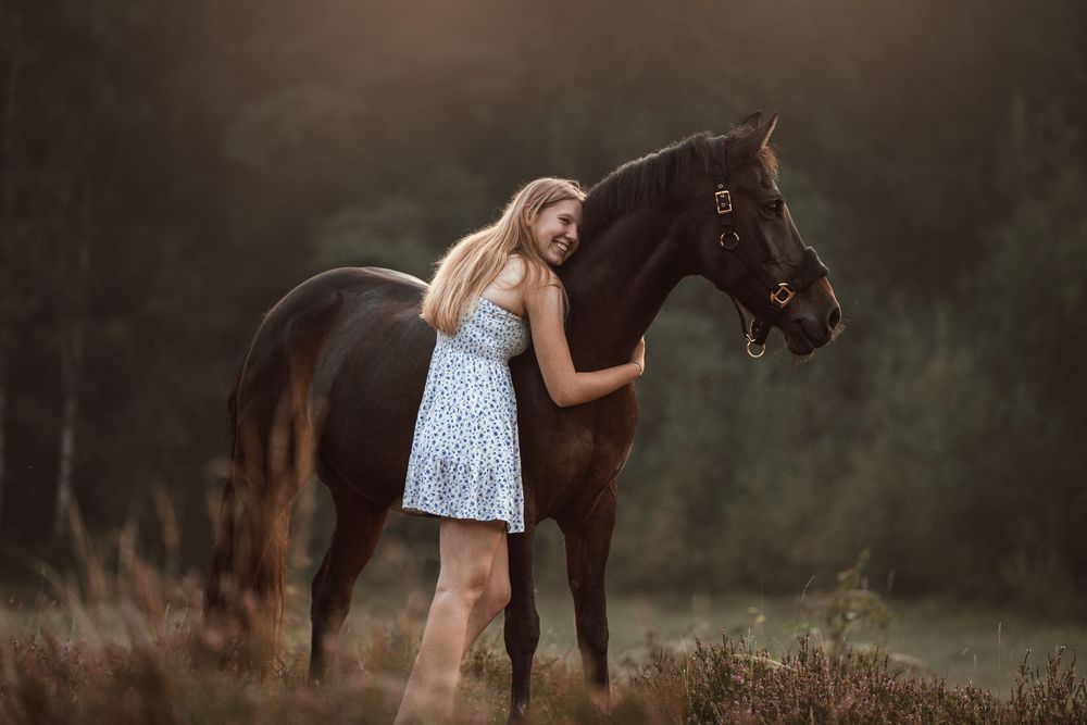 A blonde woman in a light blue dress hugs a dark brown horse in a moody, softly lit forest clearing.
