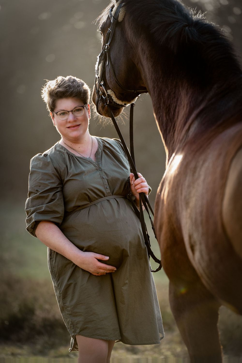 A smiling pregnant woman with glasses holds her baby bump while standing next to a brown horse in warm, hazy sunlight.