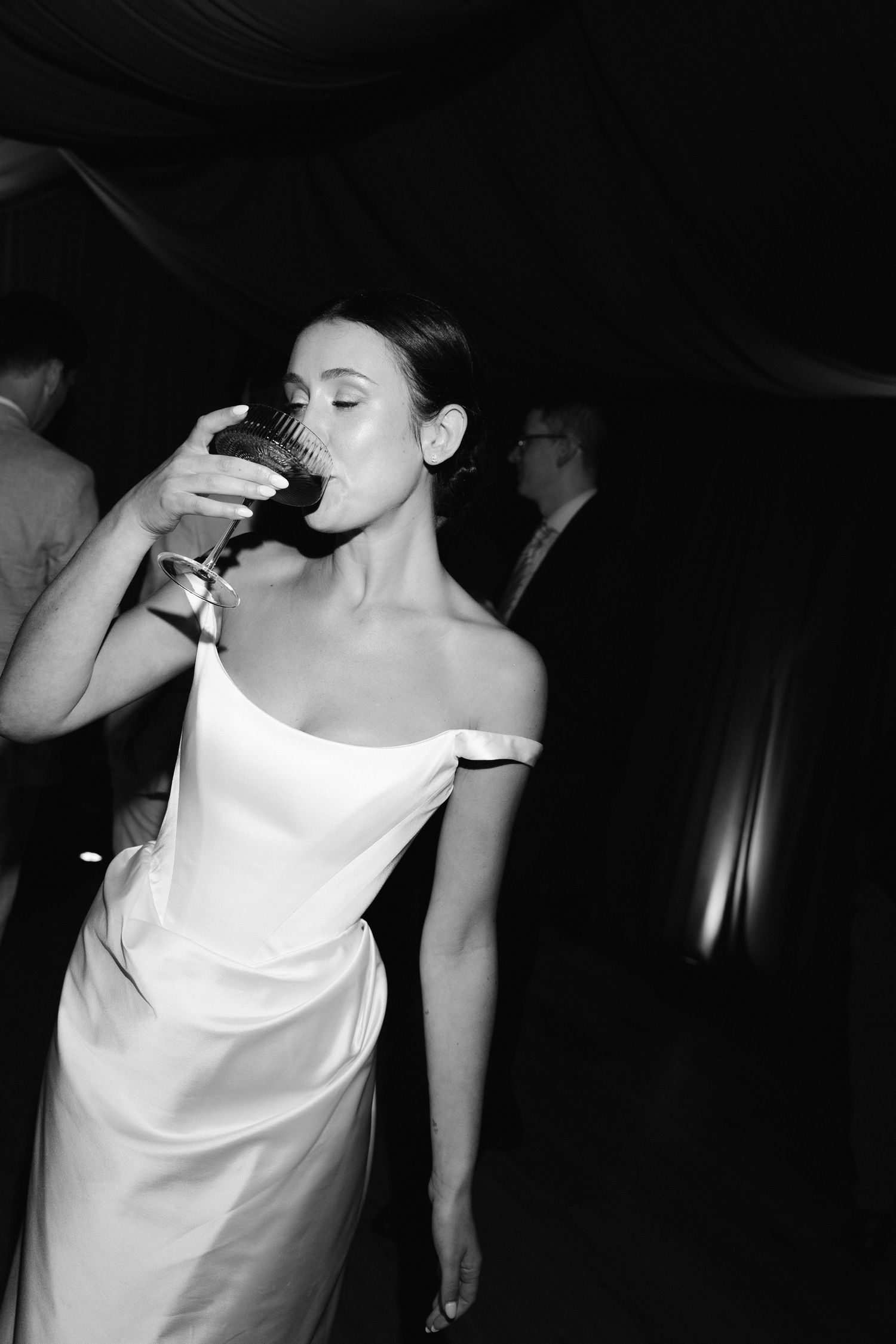 Black and white photo of a bride in an off-shoulder gown drinking wine at her wedding reception.