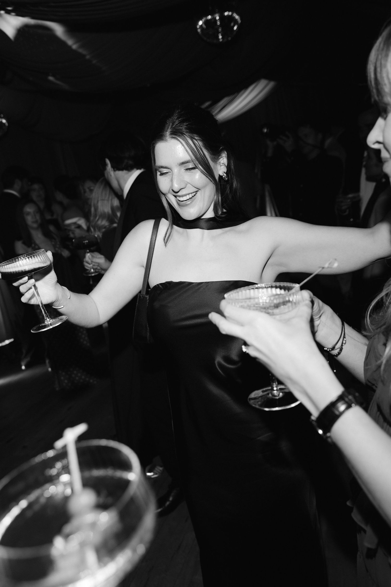 Black and white photo of a smiling woman in a black dress holding a cocktail at a lively nightclub event.