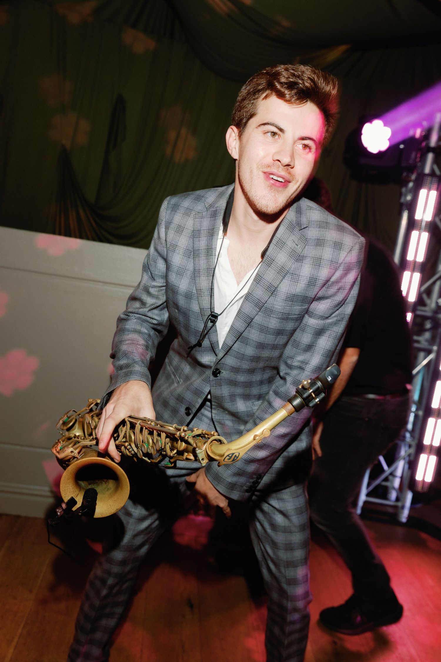 Young man in plaid suit playing saxophone at an indoor event with colorful stage lighting.
