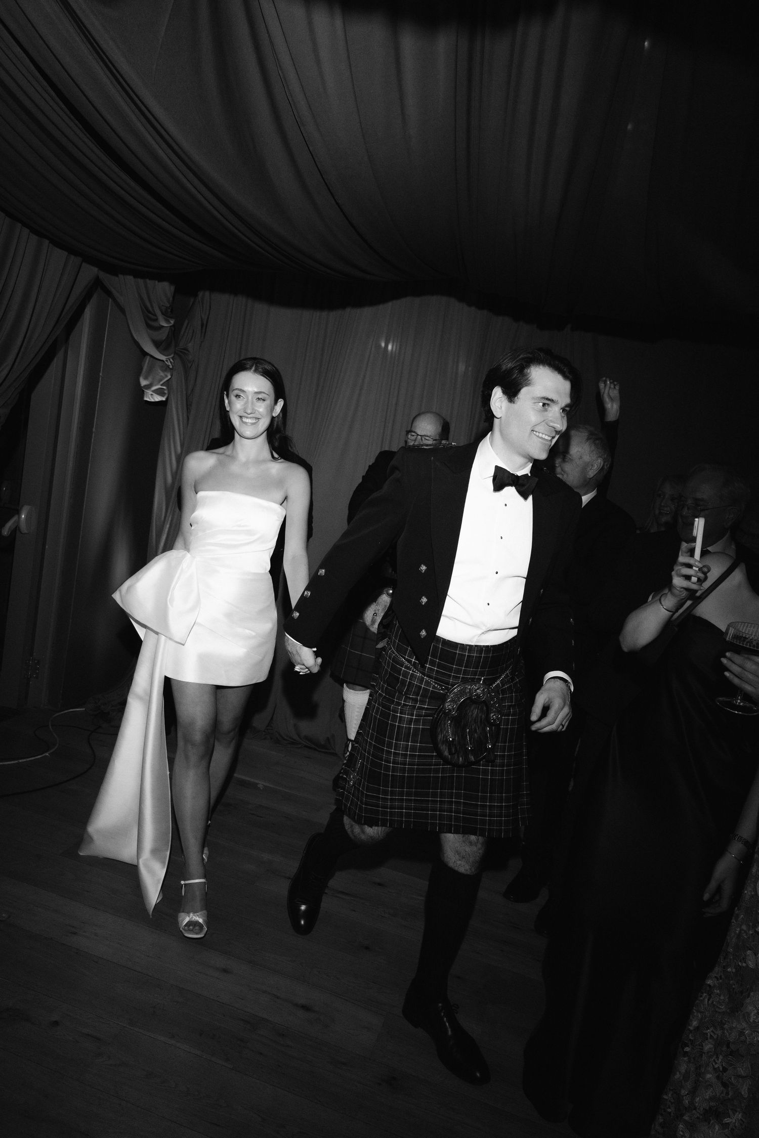 Black and white photo of bride in mini dress and groom in Scottish kilt walking at wedding reception.