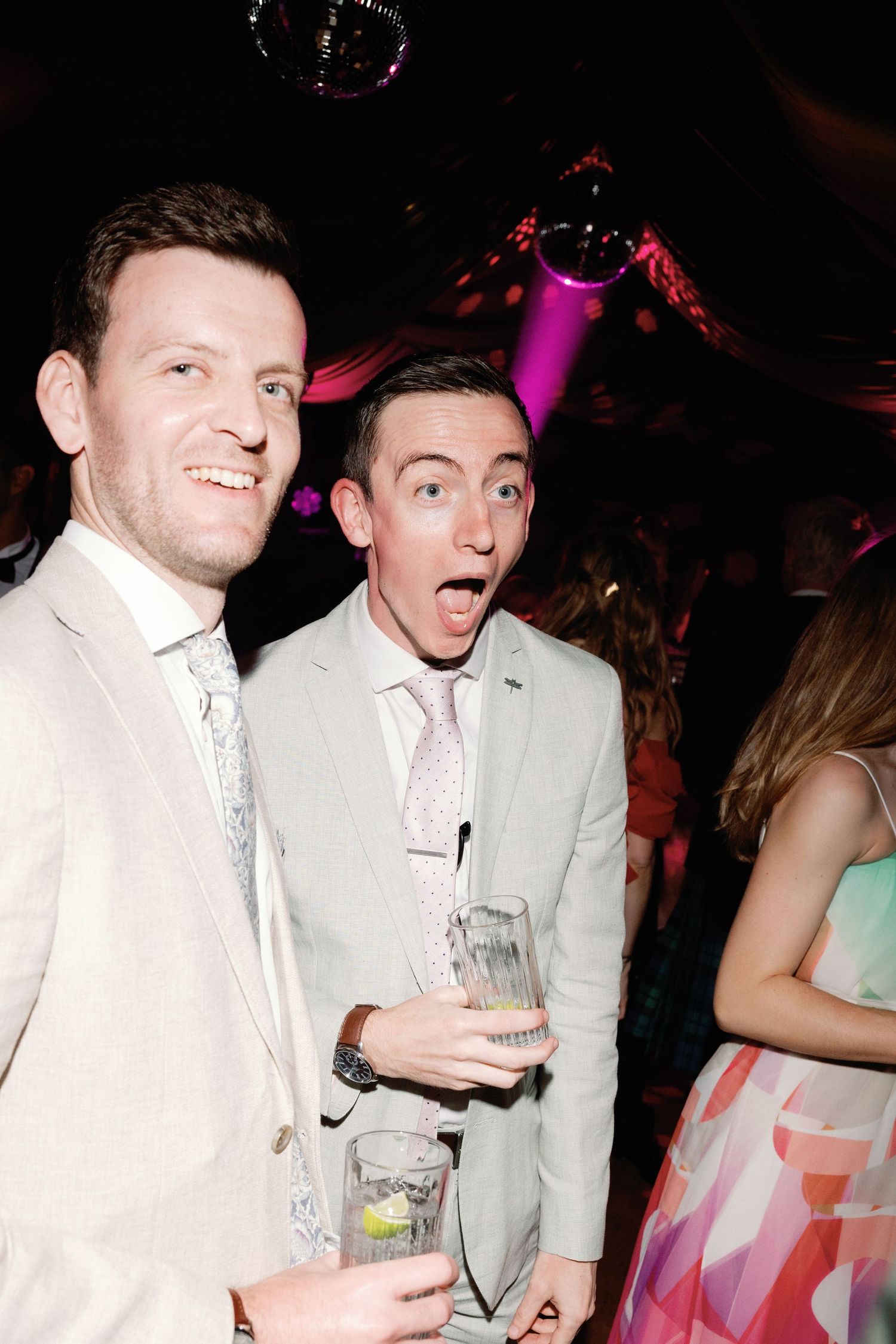 Two men in light suits smile and hold drinks at a lively wedding reception party.
