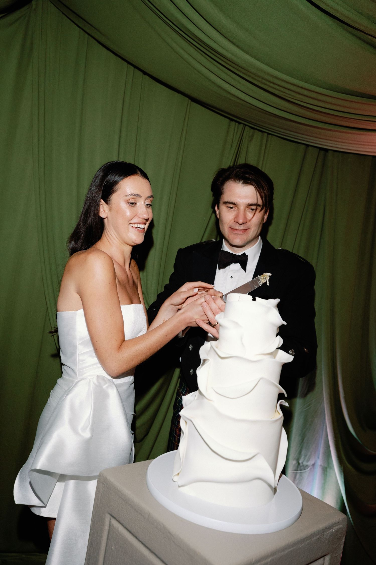 Bride and groom cut elegant white tiered wedding cake against green draped backdrop.