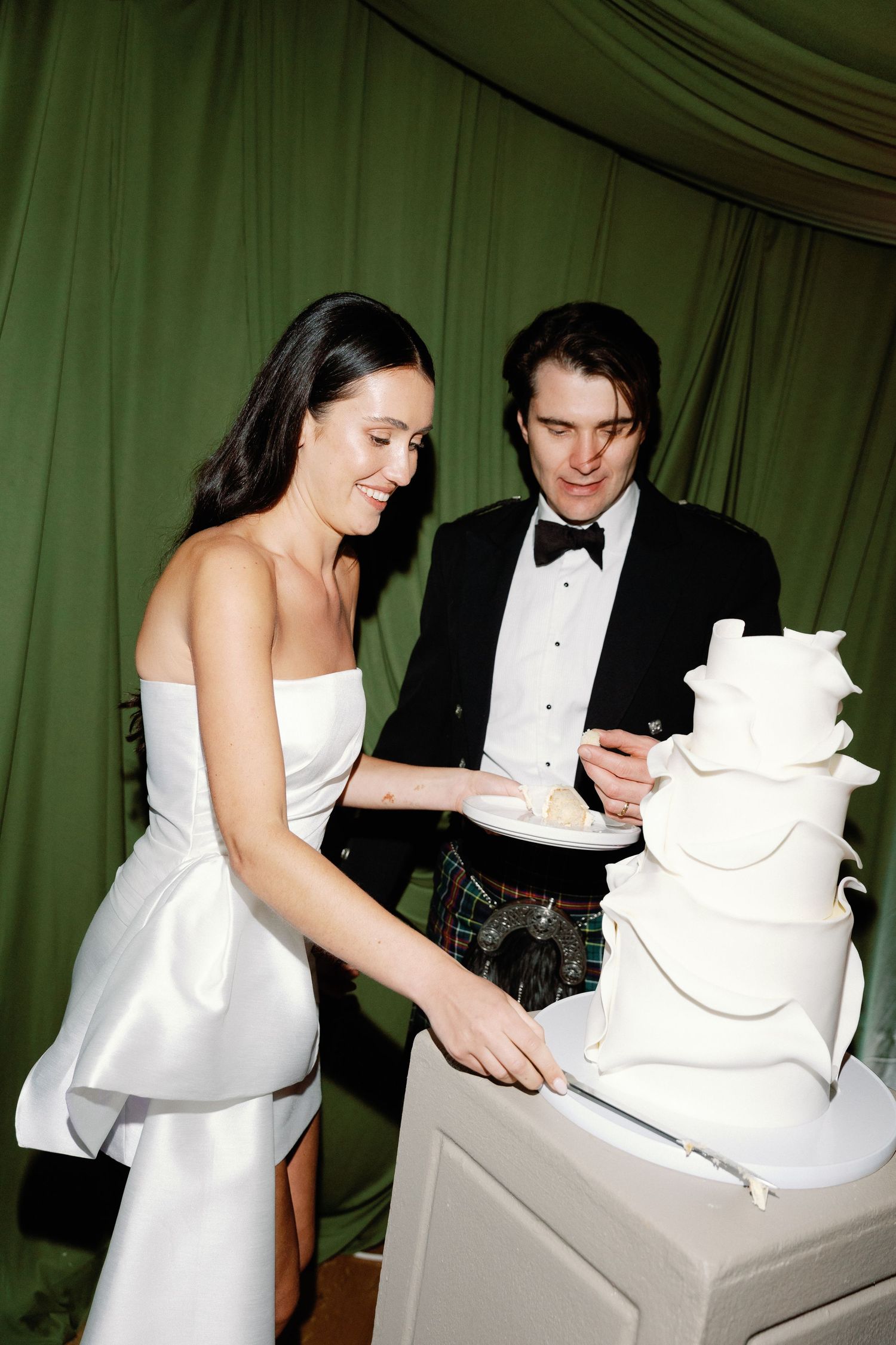 Bride in strapless gown and groom in kilt serve wedding cake at elegant reception.