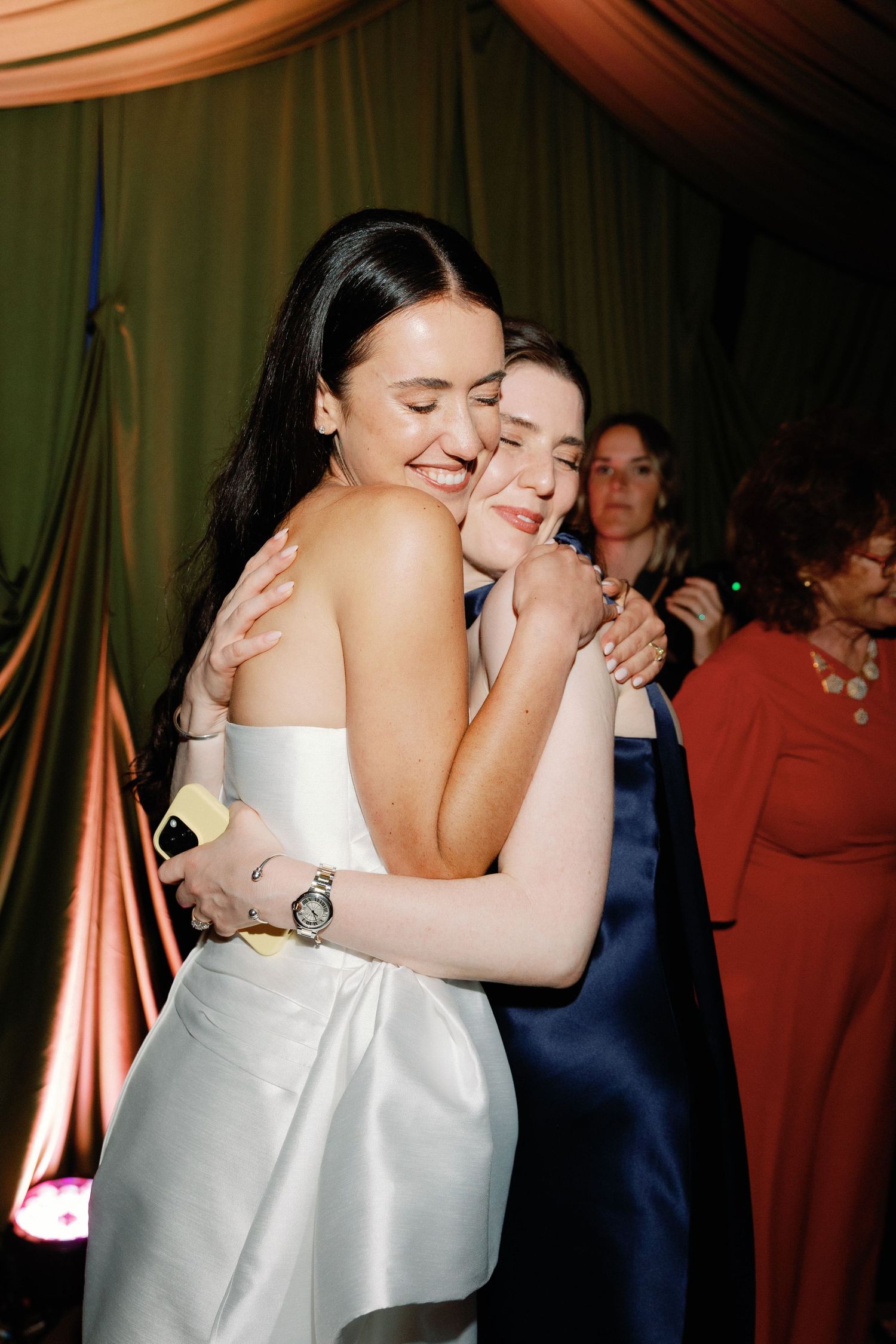 Two women in elegant gowns share a warm hug at a formal event, smiling joyfully.