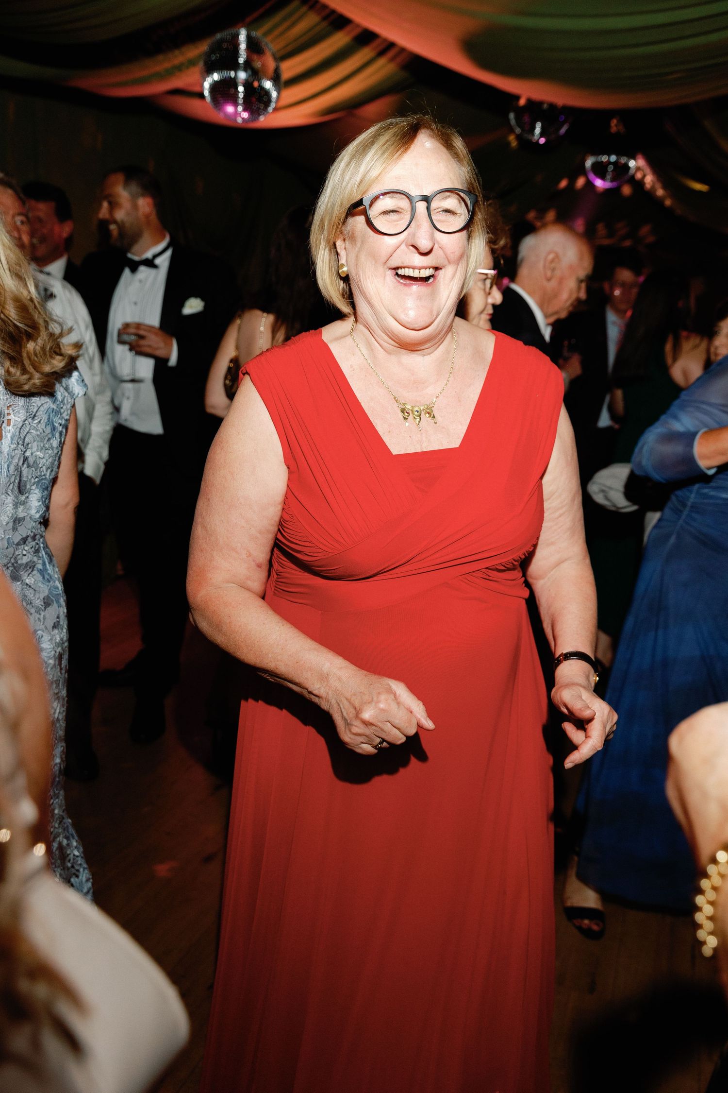 A joyful older woman in a red dress and glasses laughs while dancing at an elegant evening event.
