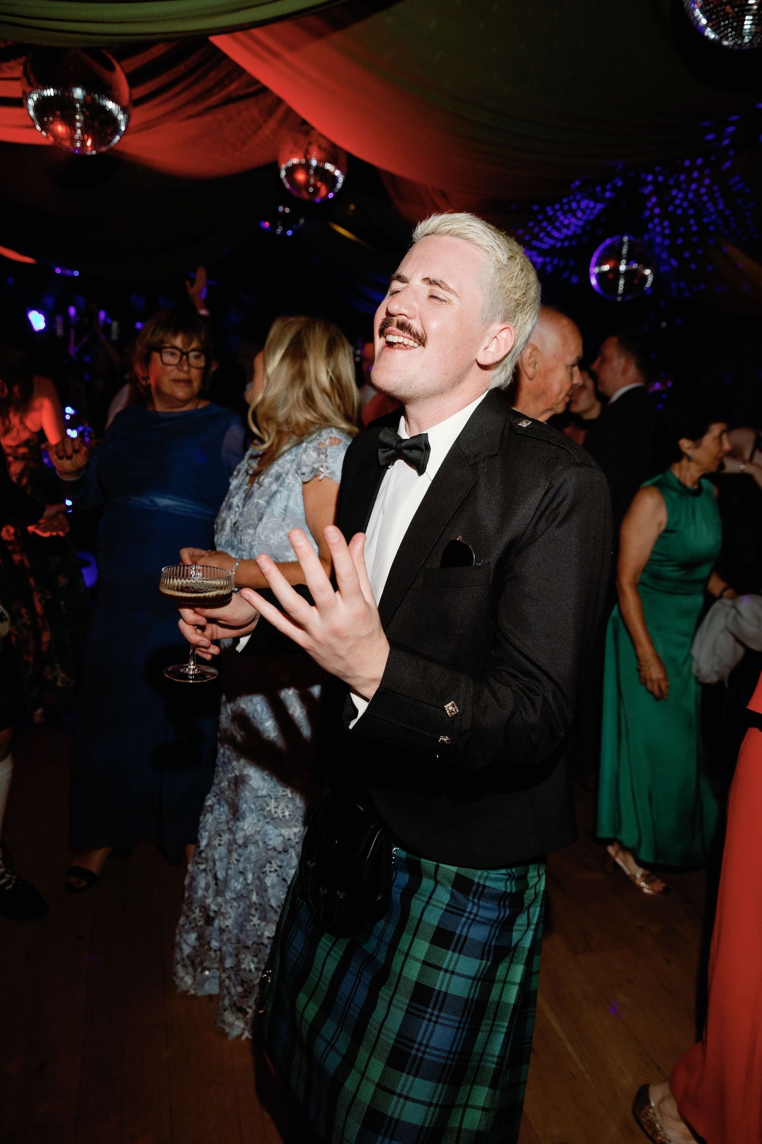 A laughing man in a kilt and black tuxedo jacket holds a drink at a formal Scottish ceilidh dance event.