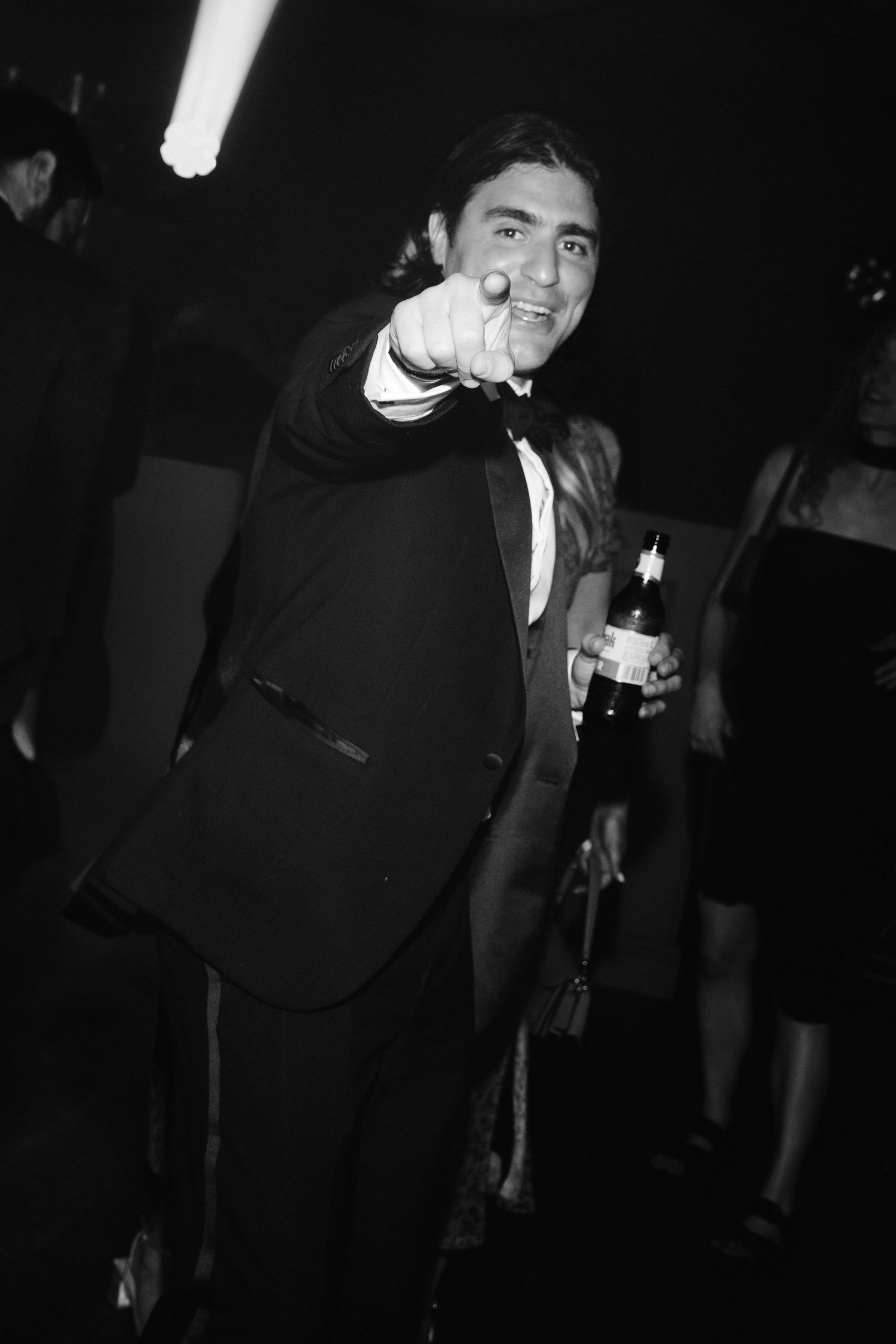 Man in tuxedo pointing at camera while holding a bottle at a formal party, black and white photo.