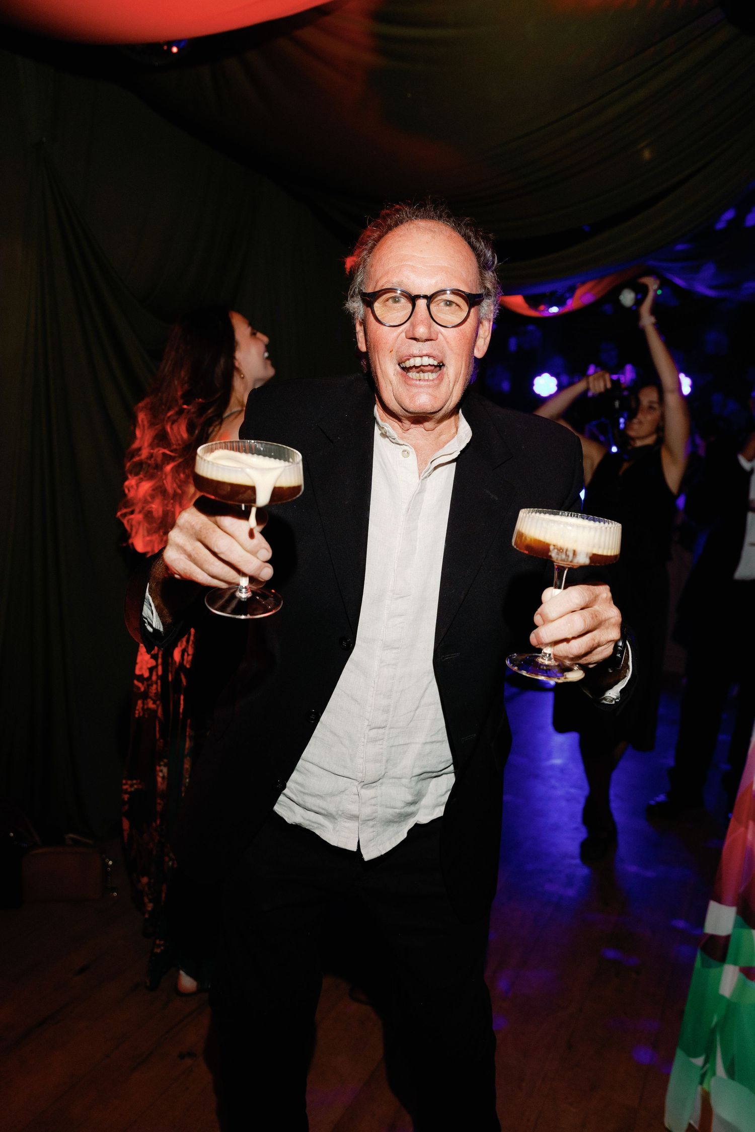 Man in black suit and glasses smiles while holding two espresso martini cocktails at a lively party.