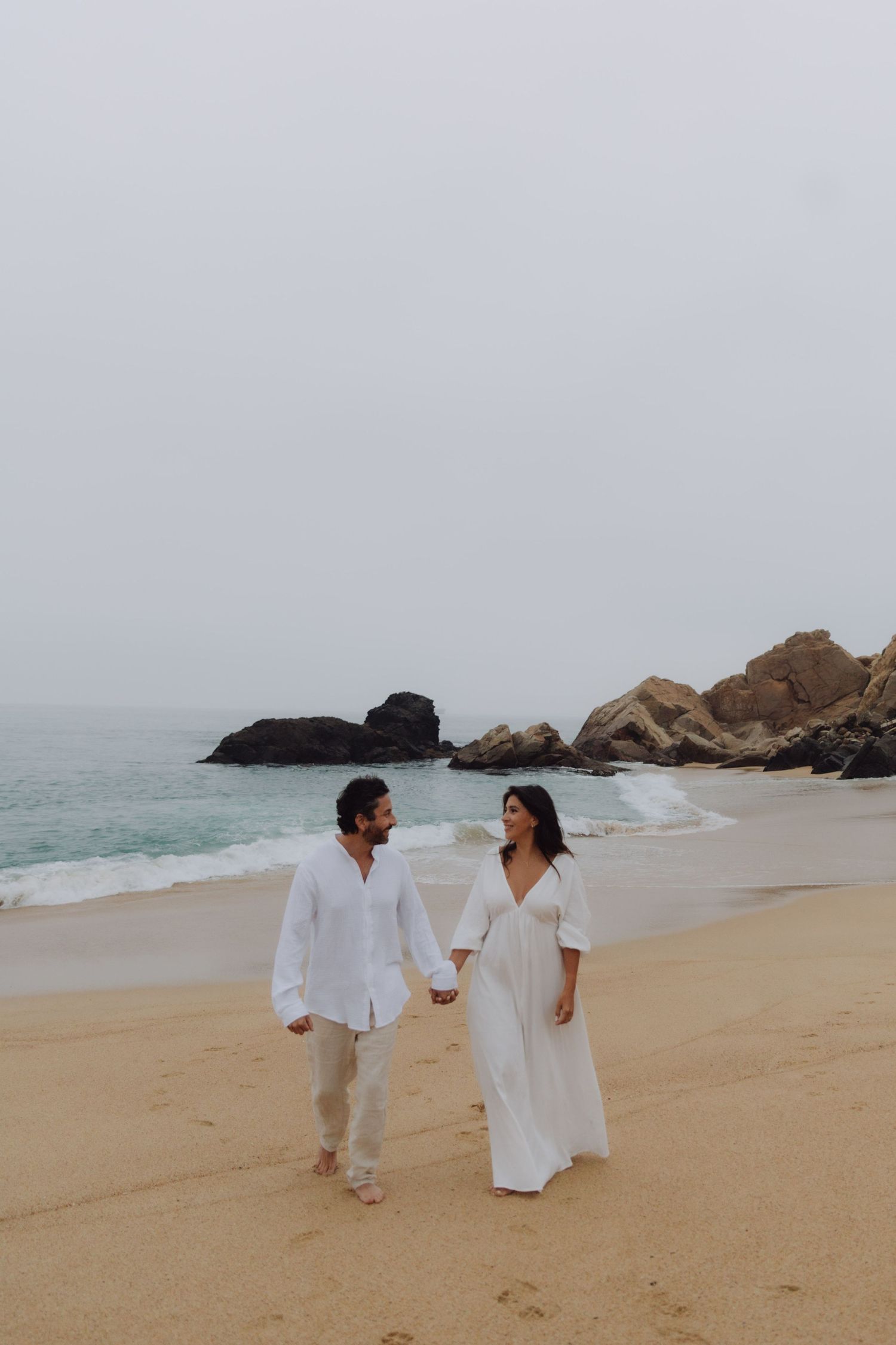Couple in white outfits holding hands walking on sandy beach with rocky shore and ocean waves behind them.