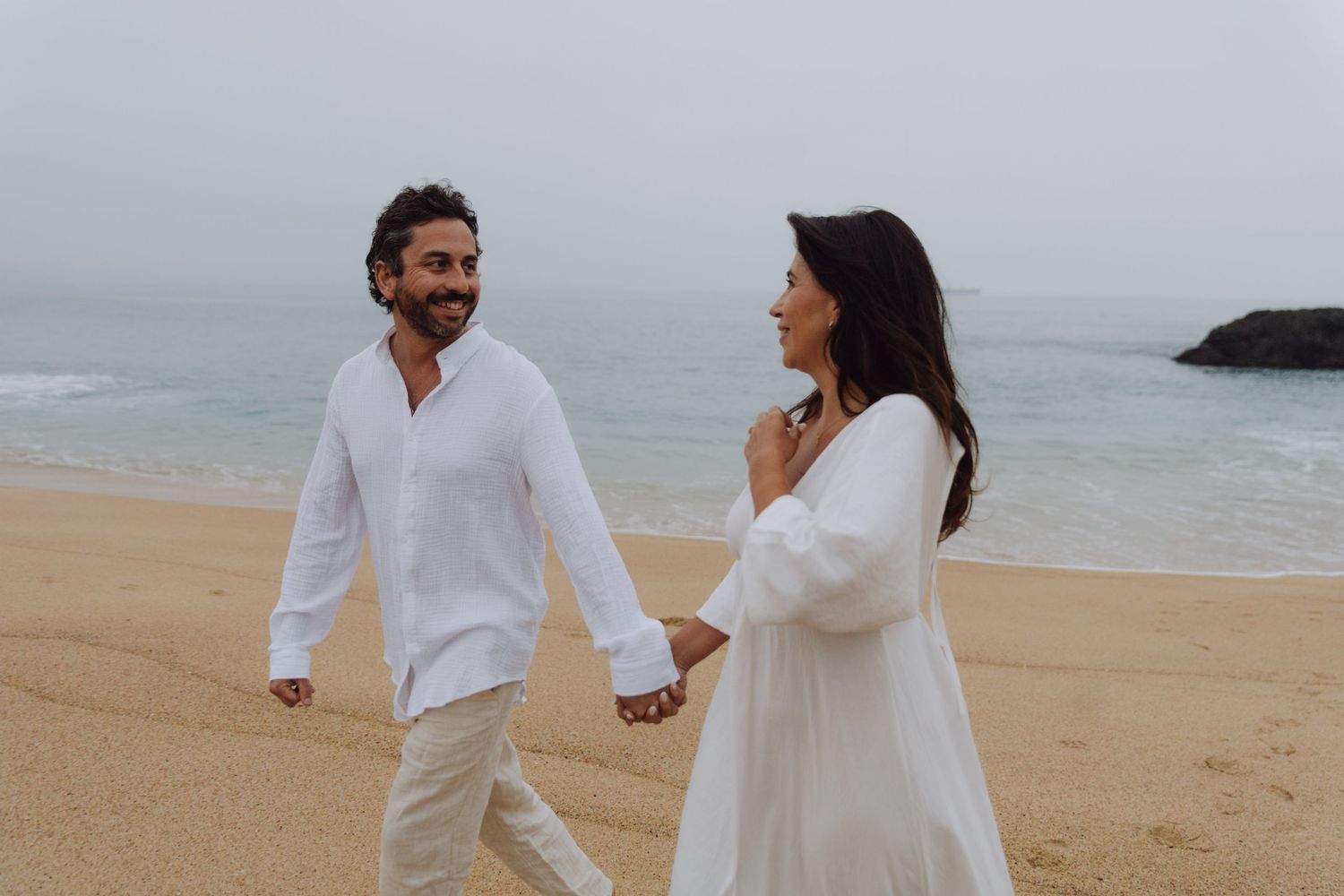 Smiling couple in white clothing holding hands strolling along sandy beach with ocean and rocks in background.