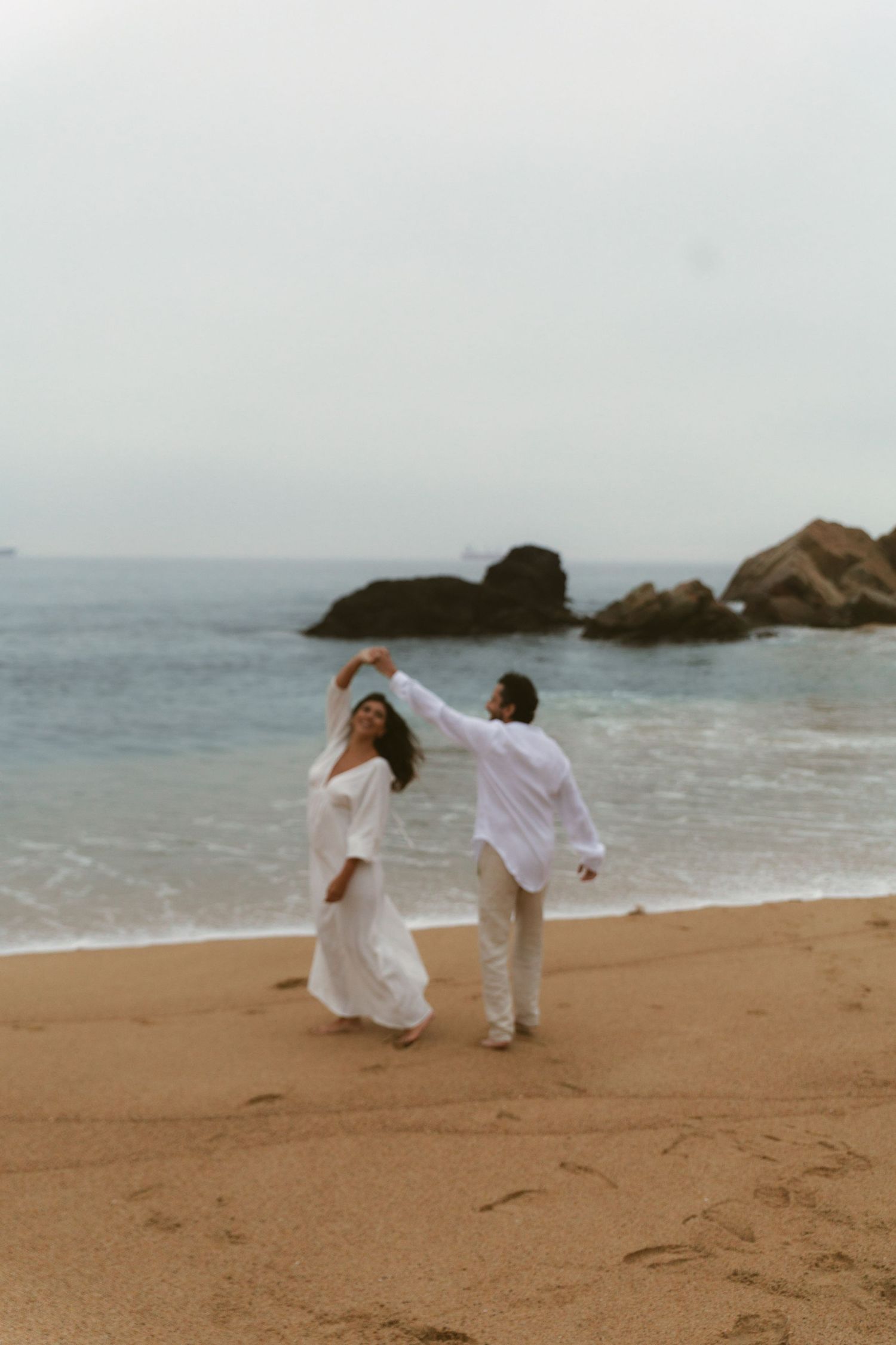 Couple in white outfits dancing on sandy beach with ocean waves and large rocks in the background.