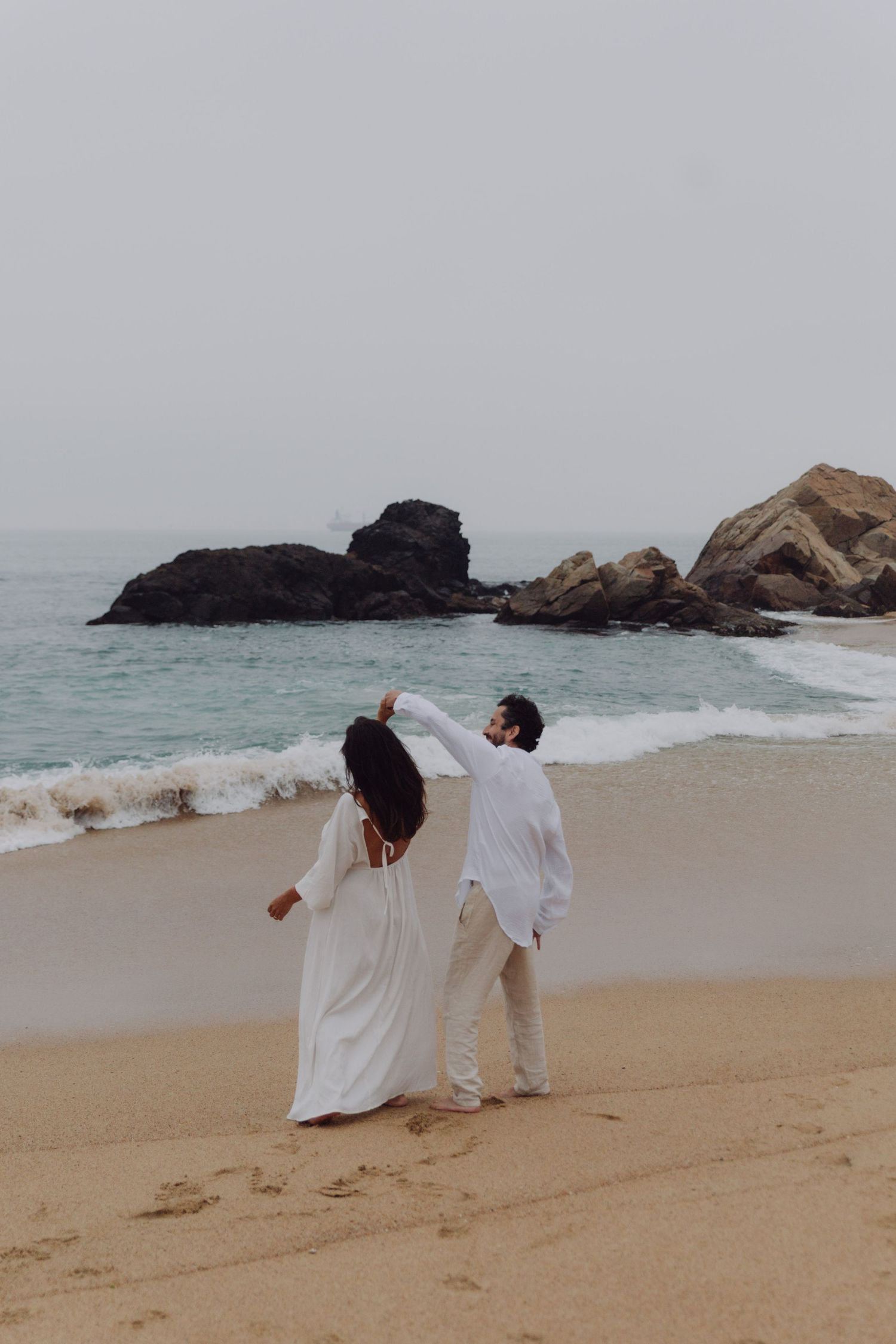 Couple in white outfits dancing on sandy beach with ocean waves and dramatic rock formations in background.